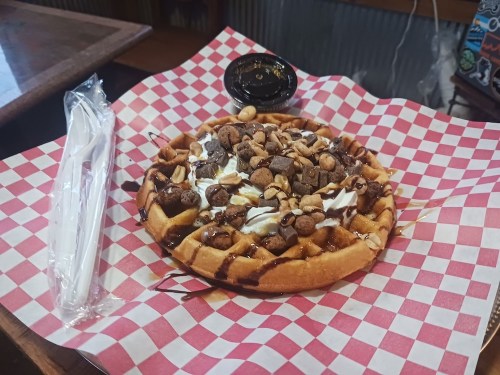 A waffle topped with whipped cream, chocolate drizzle, and nuts, served on a red and white checkered paper with utensils.