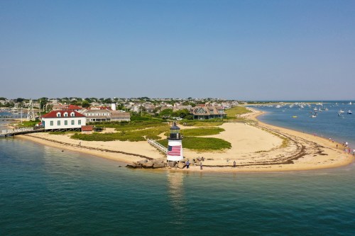 A scenic view of a lighthouse on a sandy shore, surrounded by water and boats, with a coastal town in the background.