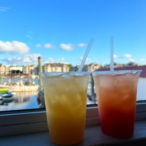 Two colorful drinks in clear cups sit on a windowsill, with a marina and blue sky in the background.