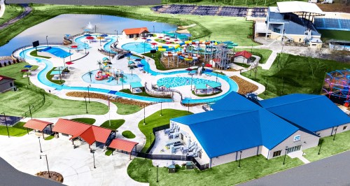 Aerial view of a vibrant water park featuring pools, slides, and play areas, surrounded by green lawns and a pond.