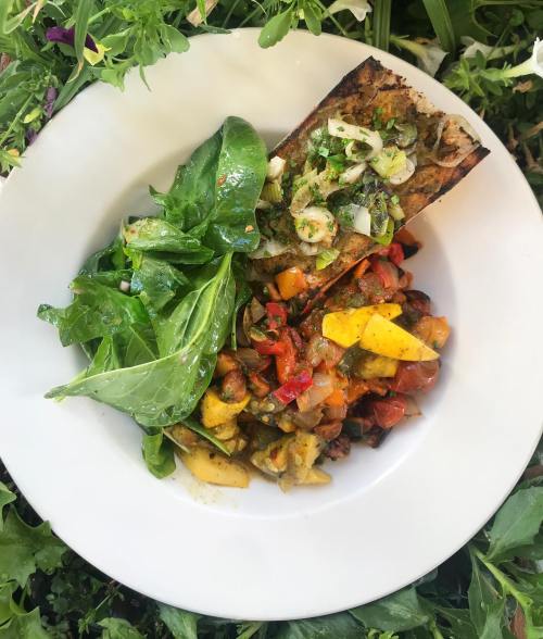 A vibrant bowl of grilled vegetables, mango, and sautéed greens, garnished with herbs, set against a floral backdrop.