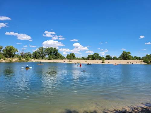 A serene river scene with people kayaking and relaxing on a sandy shore under a bright blue sky with fluffy clouds.