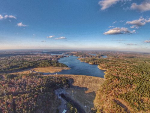 Aerial view of a serene lake surrounded by forests and rolling hills under a clear blue sky with scattered clouds.