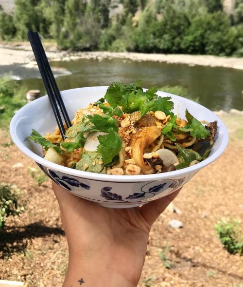 A hand holds a bowl of noodles topped with herbs and peanuts, with a river and greenery in the background.