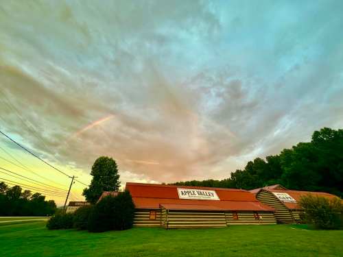 A scenic view of log buildings with "Apple Valley" signs, under a colorful sky with clouds and a hint of a rainbow.