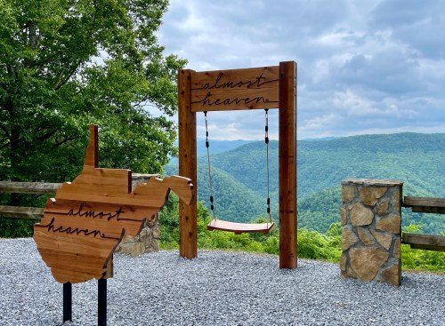 A wooden swing and sign reading "almost heaven" overlook a lush green valley under a cloudy sky in West Virginia.