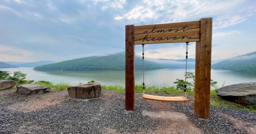 A wooden swing labeled "almost heaven" overlooks a serene lake surrounded by lush green hills under a cloudy sky.