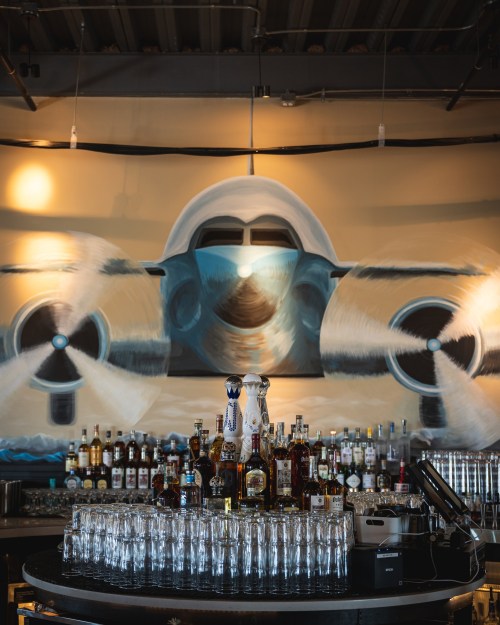 A bar with a variety of bottles and glasses, featuring a large mural of an airplane on the wall behind it.