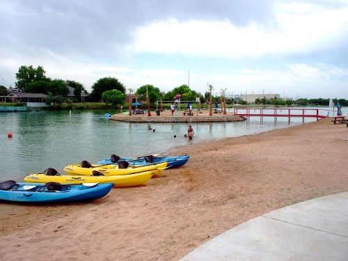 A sandy beach with colorful kayaks by a calm lake, people swimming, and a wooden dock in the background under cloudy skies.