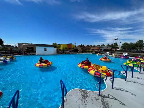 Colorful bumper boats float in a blue pool under a clear sky, with people enjoying a sunny day at an amusement park.
