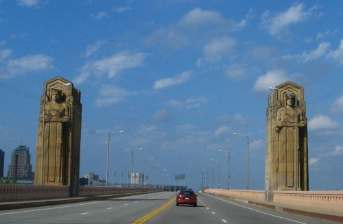 A view of a bridge with tall, decorative stone pillars and a blue sky filled with clouds. A car drives on the road.