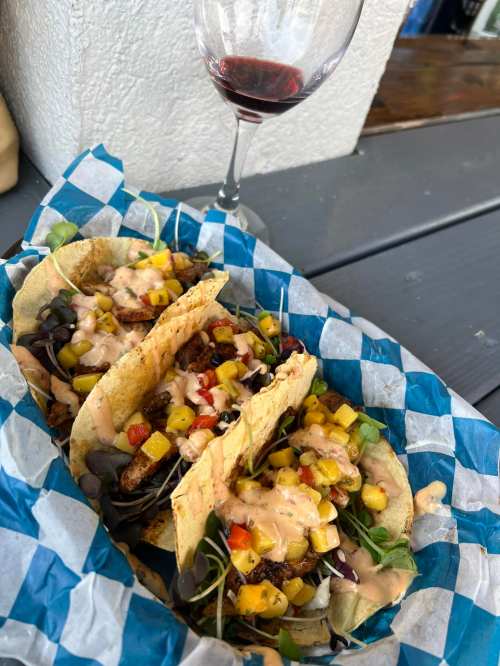 Three tacos filled with colorful toppings, drizzled with sauce, served on a blue checkered plate next to a glass of red wine.