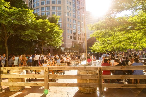 A lively outdoor gathering at Trillium Brewing, with people enjoying drinks and sunshine among trees and buildings.