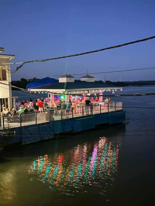A lively riverside bar at dusk, with colorful lights reflecting on the water and people enjoying the atmosphere.