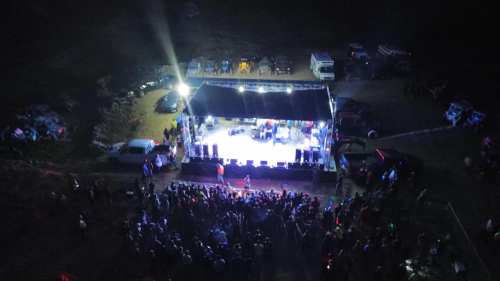Aerial view of a concert stage with bright lights and a large crowd gathered in front, surrounded by parked cars.