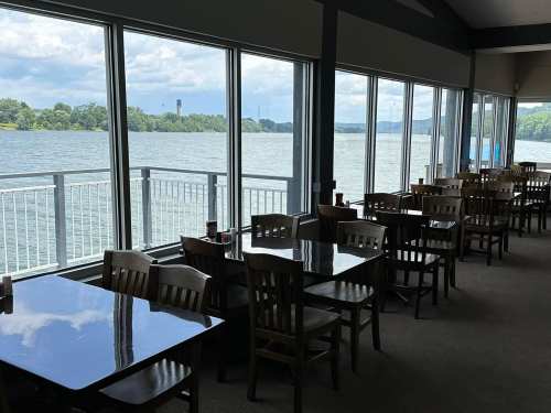 A dining area with wooden tables and chairs, overlooking a lake through large windows.