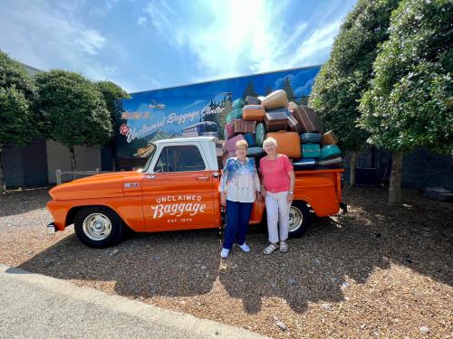 Two women stand beside an orange vintage truck loaded with colorful suitcases, in front of a mural.