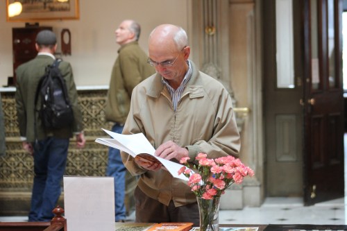 An older man in a beige jacket reads a brochure at a table with flowers in a public space.
