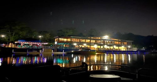 A lakeside restaurant illuminated at night, reflecting lights on the water, with people enjoying the outdoor area.