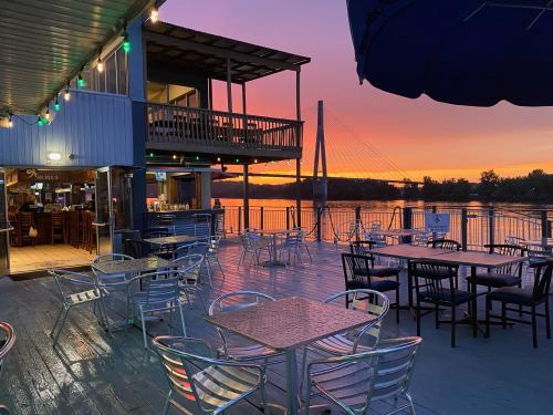 A riverside restaurant at sunset, featuring tables and chairs with a colorful sky and a bridge in the background.