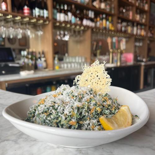 A bowl of green salad topped with grated cheese, a lemon wedge, and a crispy garnish, set against a bar background.