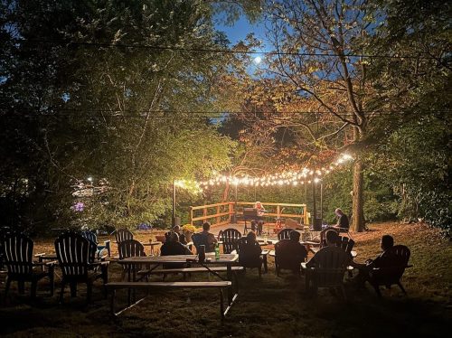 A cozy outdoor gathering at night, with string lights illuminating a stage and people seated in Adirondack chairs.