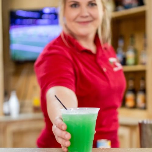 A woman in a red shirt smiles while holding a green cocktail with a straw, set against a blurred background.