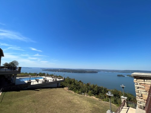 A scenic view of a lake surrounded by greenery under a clear blue sky, with a pool area in the foreground.