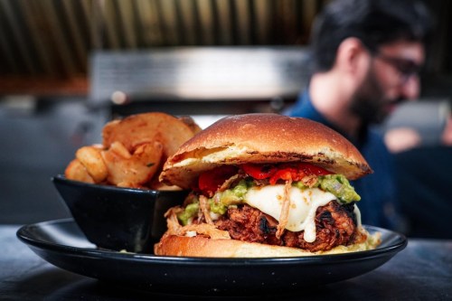 A close-up of a hearty burger topped with guacamole, peppers, and cheese, served with a side of crispy potatoes.