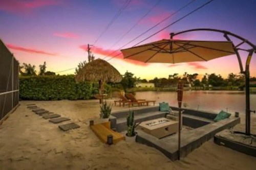 A serene beach scene with a fire pit, lounge chairs, and a thatched umbrella under a colorful sunset sky.
