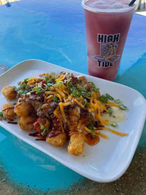 A plate of loaded tater tots topped with cheese, green onions, and barbecue sauce, alongside a colorful drink.