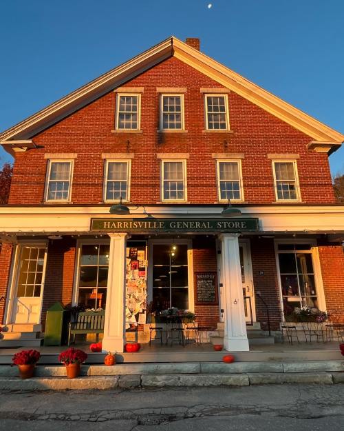 A brick building with a sign reading "Harrisville General Store," featuring pumpkins and flowers at the entrance.