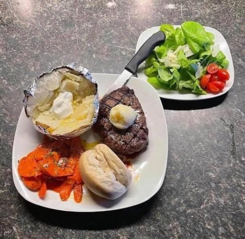 A plate with grilled steak, baked potato with sour cream, carrots, a roll, and a side salad with cherry tomatoes.