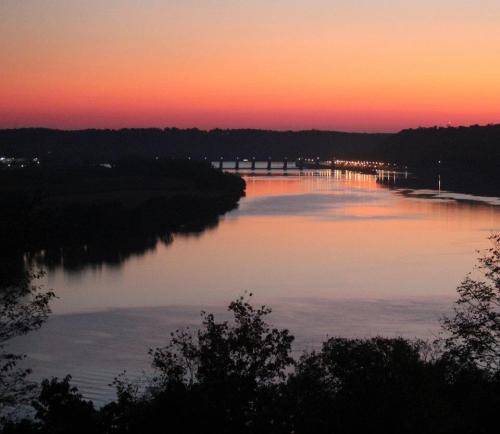 A serene river at sunset, reflecting vibrant colors in the water with a bridge and trees in the foreground.