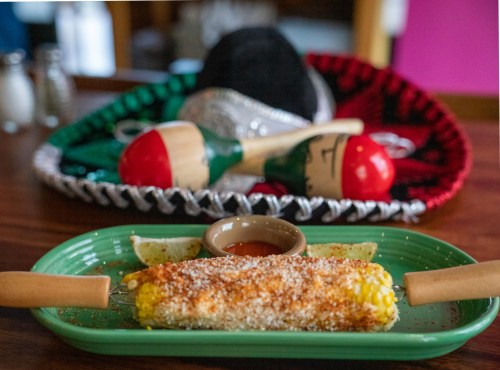 A plate of elote topped with cheese and sauce, with maracas and sombreros in the background.