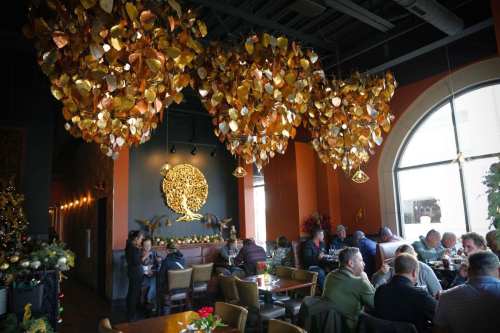 A cozy restaurant interior with golden leaf decorations, diners at tables, and a large window letting in natural light.