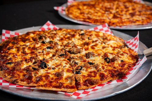 Two pizzas on metal trays, topped with cheese, sausage, and olives, on a black table with red checkered paper.