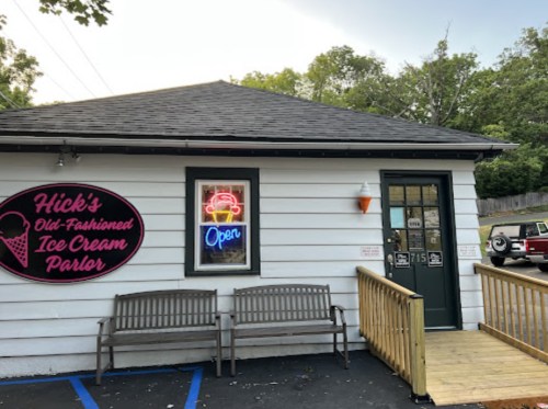 A quaint ice cream parlor with a neon "Open" sign and a welcoming porch, surrounded by trees.