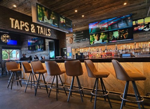 A modern bar with high stools, illuminated taps, and multiple TVs displaying sports in a rustic setting.