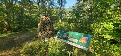 A stone cairn surrounded by trees, with an informational sign about the area in a lush, green forest.