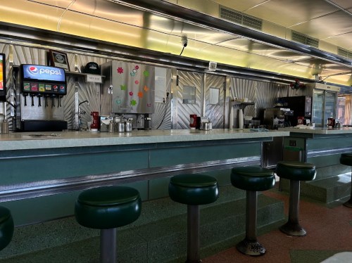 A retro diner interior featuring a long counter with green stools, soda fountain, and colorful decorations on the wall.