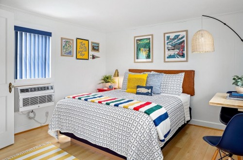 Cozy bedroom with a colorful quilt, wall art, and a small desk, featuring blue accents and natural light.