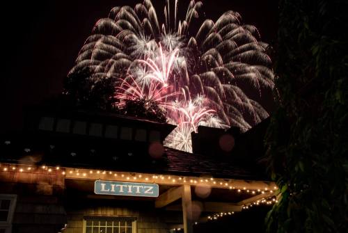 Colorful fireworks burst in the night sky above a building with a sign reading "Lititz," illuminated by string lights.