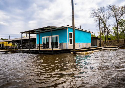 A bright blue house on stilts surrounded by water, with a cloudy sky in the background.