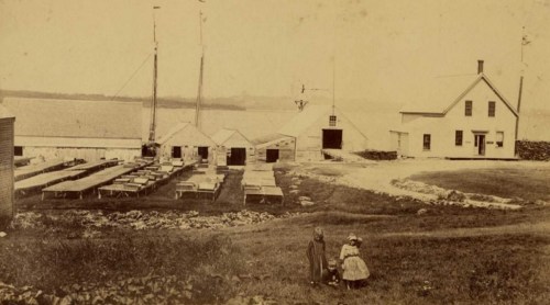 Historic black-and-white photo of two girls in front of a coastal building with boats and storage sheds in the background.
