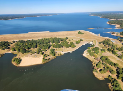 Aerial view of a serene lake with sandy beaches, green trees, and a dam in the distance under a clear blue sky.