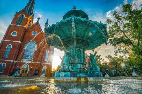 A decorative fountain with water spouting, set against a backdrop of a historic brick church and a vibrant sky.