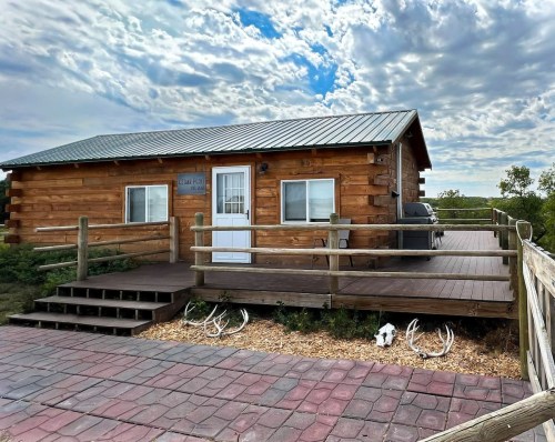 A rustic wooden cabin with a metal roof, surrounded by greenery and featuring a spacious deck and stone pathway.