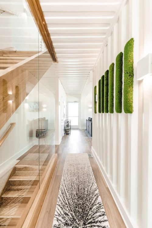 A modern hallway with wooden flooring, a patterned rug, and green wall art, featuring a staircase and natural light.