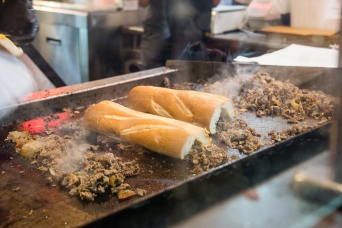 Two toasted hoagie rolls filled with sizzling, chopped beef on a griddle, surrounded by steam and cooking ingredients.
